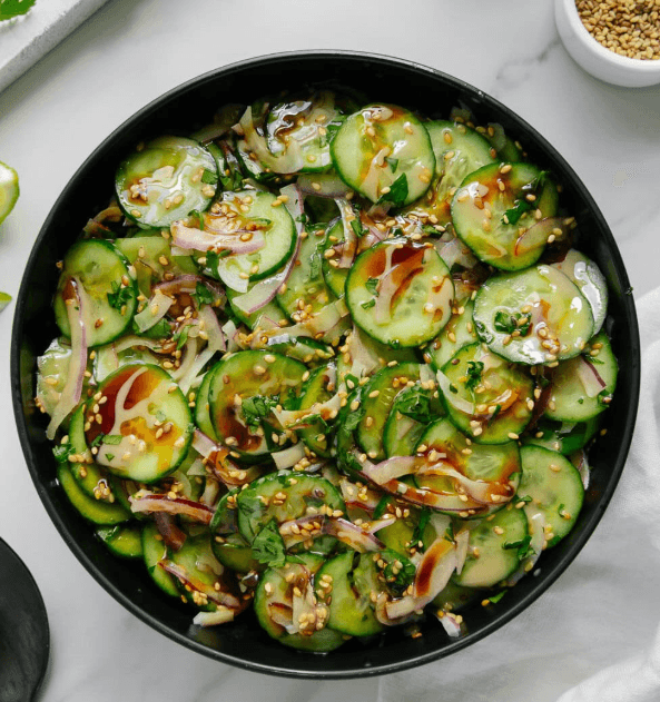 Sliced cucumber salad with red onions, sesame seeds, and herbs in a black bowl.